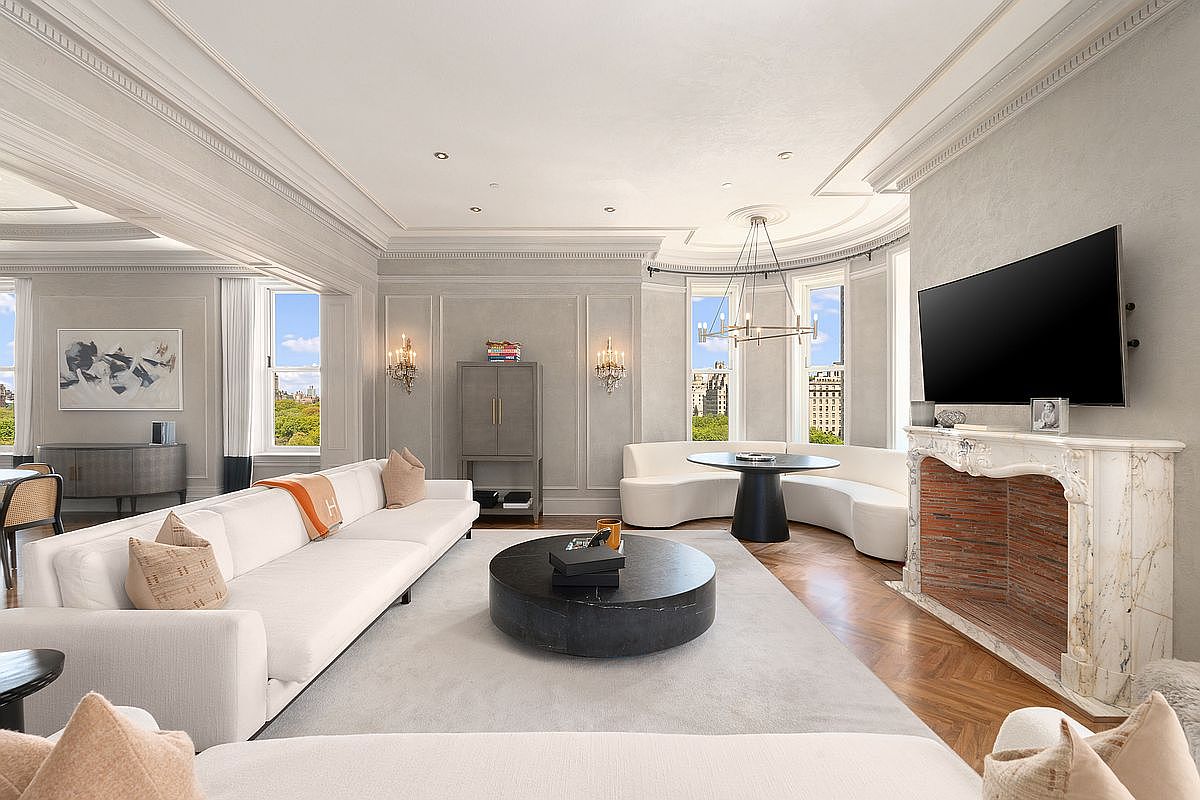 This elegant, expansive living room features a sophisticated neutral color palette, high ceilings with intricate crown molding, and a herringbone-patterned wood floor. The space is anchored by a large, circular dark stone coffee table and a long, white sofa, while a curved banquette seating area sits near the tall windows that offer views of the city. A prominent fireplace with a decorative stone surround and a mounted television serves as a focal point, complemented by wall sconces and a modern chandelier.