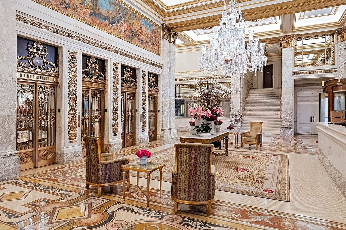 This grand, opulent lobby features intricate stone flooring with elaborate patterns, ornate gold-trimmed elevator doors, and a massive crystal chandelier suspended from a high, detailed ceiling. A central console table with a large floral arrangement serves as a focal point, surrounded by elegant upholstered armchairs and a sweeping marble staircase in the background. The space exudes a sense of historic luxury and high-end sophistication, typical of a prestigious building's entrance.