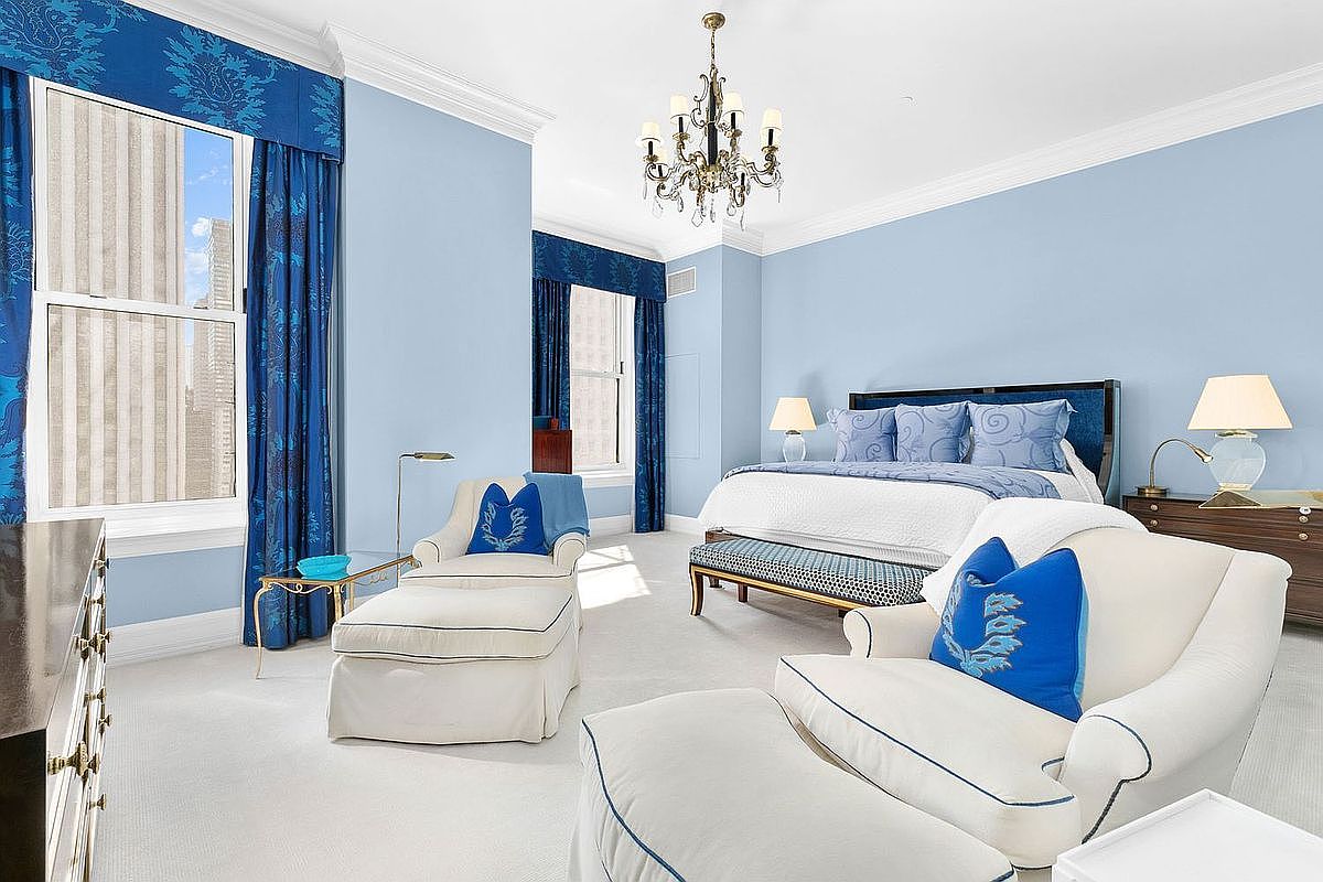 This elegant primary bedroom features a sophisticated blue and white color palette, anchored by a large bed with a dark frame and patterned accent pillows. The room is furnished with two comfortable armchairs with matching ottomans, a crystal chandelier, and tall windows dressed in vibrant blue drapery. The space offers a bright, airy atmosphere with plush carpeting and a classic, refined aesthetic.