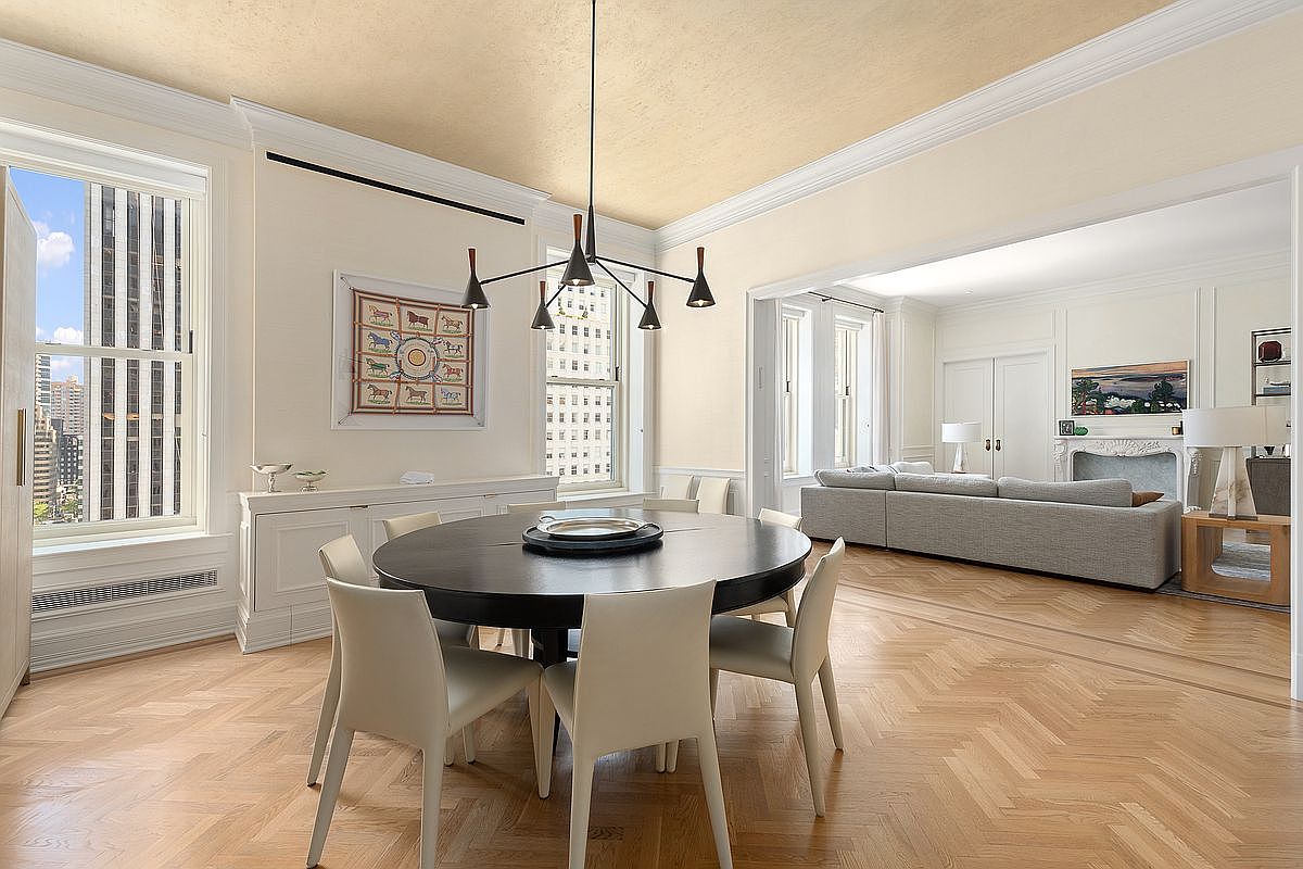 This elegant dining room features a dark circular table surrounded by six light-colored upholstered chairs, set against a backdrop of herringbone-patterned wood flooring. A modern, multi-armed chandelier serves as a focal point above the table, while a long, low-profile sideboard sits beneath a framed piece of art. The room flows seamlessly into an adjacent living area, which is anchored by a fireplace and a large gray sectional sofa, creating an open and sophisticated atmosphere.