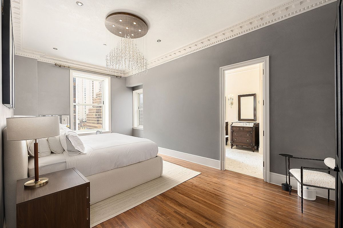 This elegant primary bedroom features a neutral color palette with soft gray walls, crown molding, and rich wood flooring. A large, plush bed serves as the focal point, complemented by a striking crystal chandelier and a modern bedside lamp. The room flows into an adjacent space through a doorway, offering a glimpse of a classic dresser and mirror, creating a sophisticated and airy atmosphere.