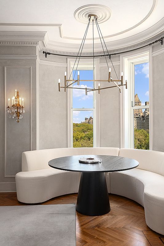 This elegant dining area features a curved, cream-colored banquette set against a semi-circular wall with tall windows overlooking a cityscape. A modern, multi-armed chandelier hangs above a dark, circular pedestal table, creating a sophisticated focal point. The room is finished with herringbone wood flooring and classic wall molding, offering a refined and airy atmosphere.
