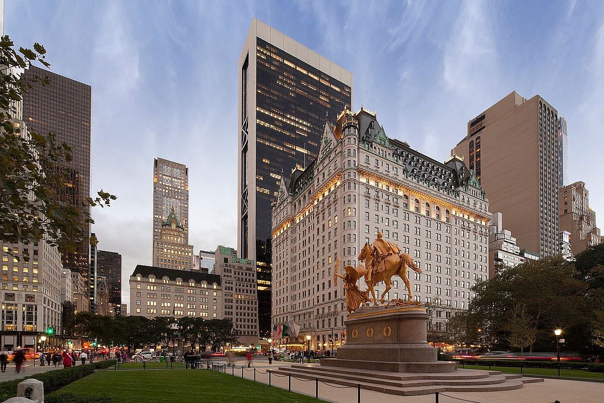 This cinematic wide-angle shot captures the iconic Plaza Hotel in New York City, showcasing its ornate French Renaissance-inspired architecture and distinctive green mansard roof. In the foreground, the General William Tecumseh Sherman Monument stands prominently within the park setting, framed by lush greenery and paved walkways. The composition highlights the contrast between the historic hotel's intricate facade and the surrounding modern skyscrapers, creating a grand urban atmosphere.