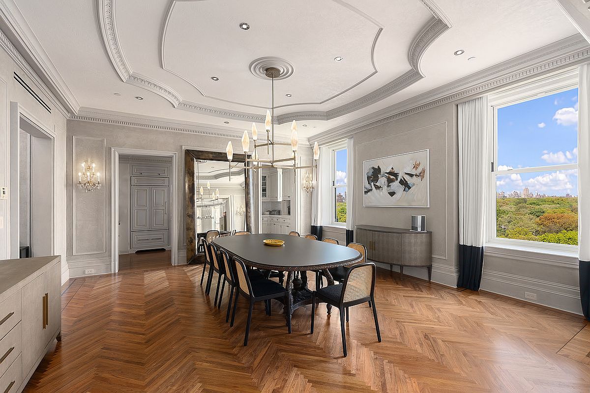 This elegant dining room features a sophisticated oval table surrounded by black chairs with woven cane backs, set atop a rich herringbone-patterned wood floor. A large, ornate chandelier serves as the focal point beneath a detailed tray ceiling, while a massive mirror reflects the space and adds depth. Tall windows on the right provide abundant natural light and a scenic view of a lush green park, creating a bright and refined atmosphere.