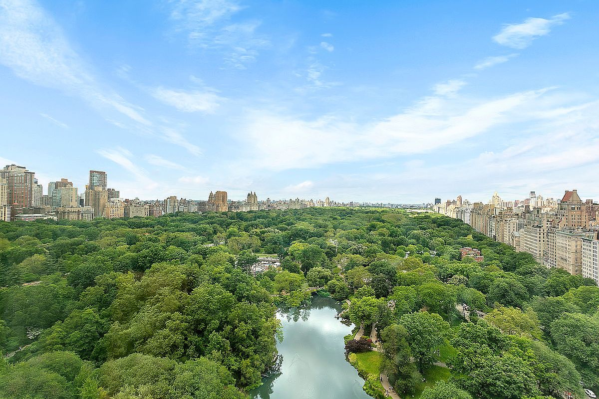 This high-angle aerial view captures a breathtaking perspective of Central Park in New York City, showcasing the lush green canopy of trees and a serene lake in the foreground. The surrounding cityscape features a dense collection of historic and modern high-rise buildings lining the park's perimeter under a bright, expansive blue sky. The composition emphasizes the scale and prestige of the location, highlighting the unique juxtaposition of urban density and natural landscape.