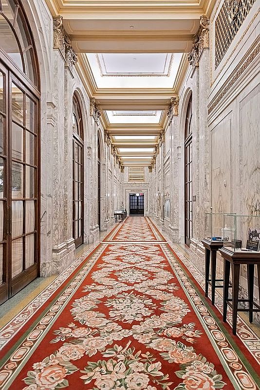 This grand, elongated hallway features polished stone walls and a striking red patterned runner rug that stretches the length of the corridor. The space is defined by tall, arched glass doors and a series of recessed ceiling panels accented with ornate gold-toned molding. The perspective is a centered, symmetrical shot looking down the hall, emphasizing the opulent and formal architectural style.