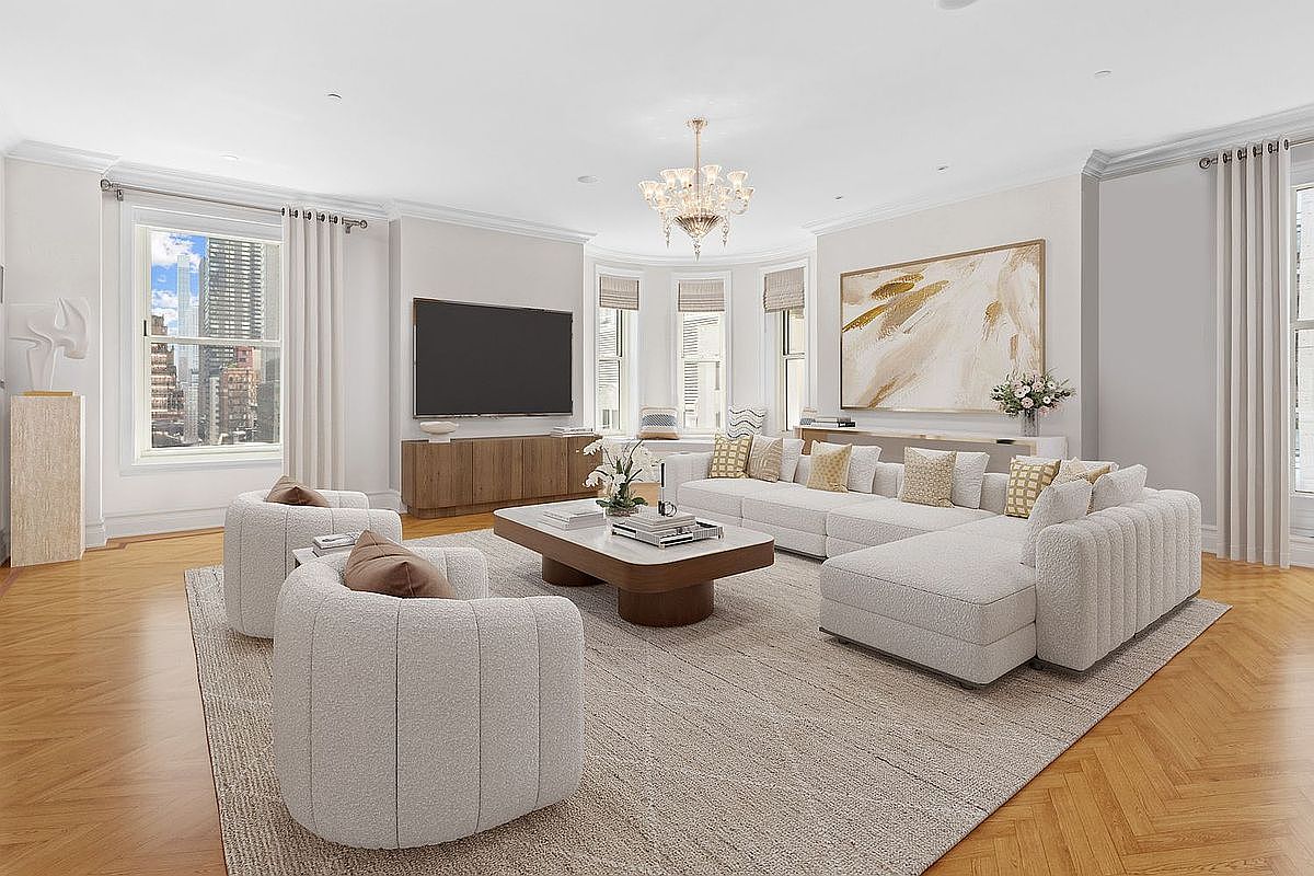 This bright and airy living room features a large, neutral-toned sectional sofa and two matching curved armchairs arranged around a low, rectangular coffee table. The space is anchored by a textured area rug over herringbone-patterned wood floors, with a large television mounted above a light wood media console. Large windows with white curtains allow for abundant natural light and offer a view of the city skyline, while a crystal chandelier adds a touch of elegance to the room.
