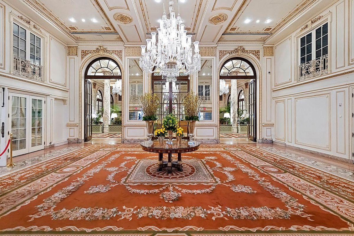 This grand, formal foyer features an expansive, ornate orange patterned rug centered beneath a massive, multi-tiered crystal chandelier. The space is defined by high ceilings with intricate crown molding, symmetrical arched doorways leading to adjacent rooms, and mirrored wall panels that enhance the sense of scale. A circular pedestal table adorned with floral arrangements serves as a focal point in this opulent, classically styled entrance hall.