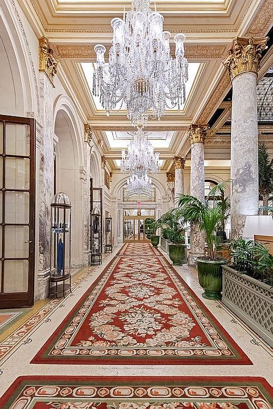 This grand, opulent hallway features a long, ornate red patterned runner leading through a series of towering stone columns. Two magnificent crystal chandeliers hang from the high, detailed ceiling, casting a warm glow over the polished stone floors and decorative wall moldings. The perspective is a centered, symmetrical shot that emphasizes the luxurious scale and classical architectural elegance of the space.