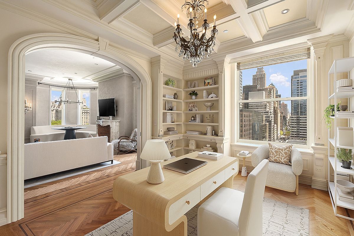 This elegant home office features a light wood desk, a modern white chair, and built-in shelving that wraps around the corner. A large window provides ample natural light and a view of the city skyline, while a crystal chandelier adds a touch of sophistication to the coffered ceiling. The room flows seamlessly through a wide arched opening into an adjacent living area, showcasing a cohesive and luxurious interior design.