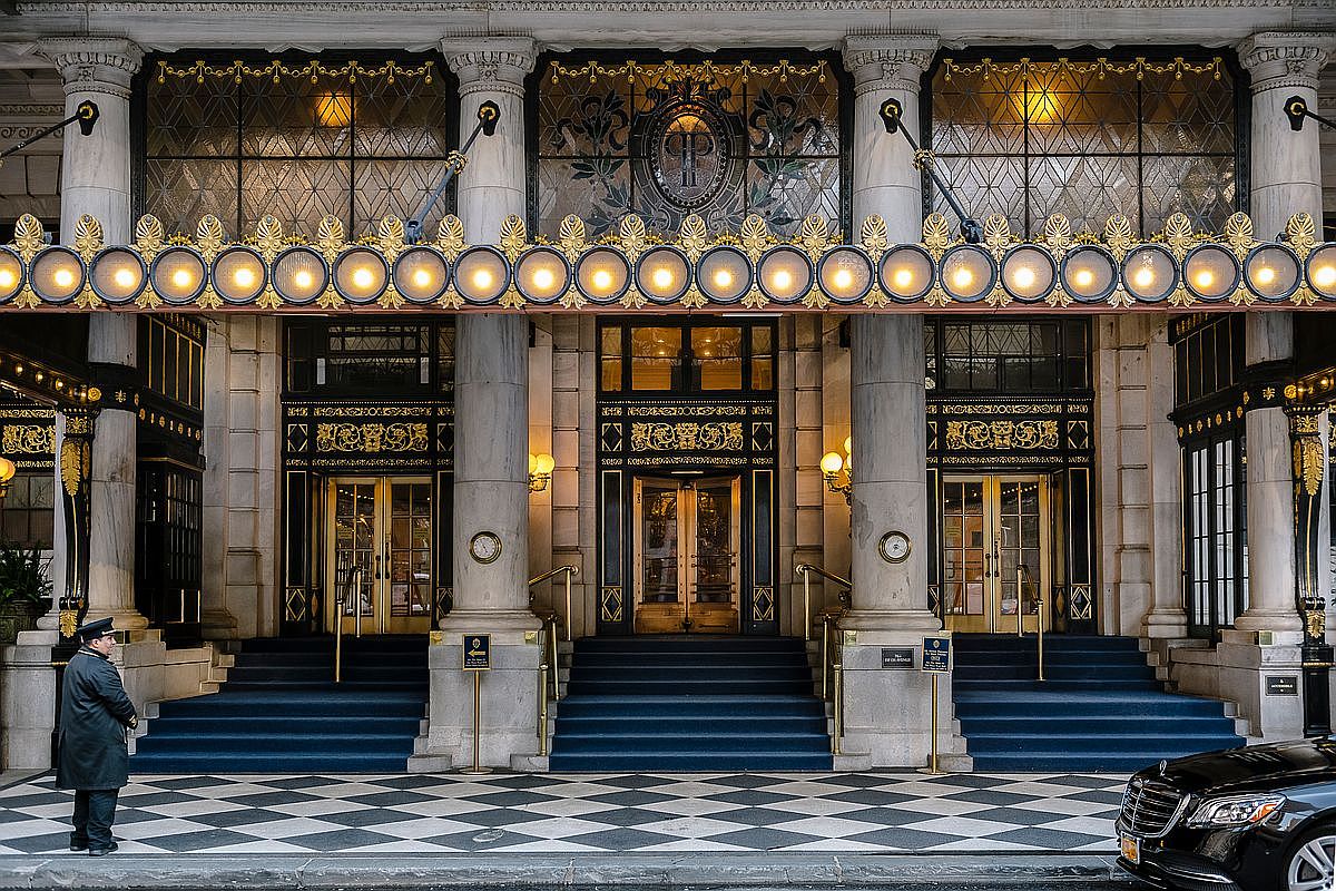 This grand, ornate entrance features a symmetrical facade with massive stone columns and a prominent marquee lined with glowing circular lights. Three sets of double doors with intricate gold-toned metalwork are accessed by wide, blue-carpeted steps, while a black-and-white checkered floor spans the foreground. A uniformed doorman stands to the left, and a luxury vehicle is partially visible on the right, emphasizing the prestigious and historic character of the building.