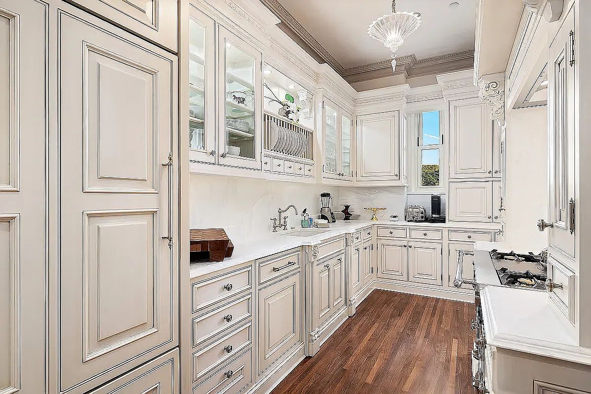 This elegant, U-shaped kitchen features classic off-white cabinetry with intricate molding and a distressed finish, paired with light-toned stone countertops. The space is equipped with high-end integrated appliances, including a paneled refrigerator and a gas range, complemented by warm wood flooring and a decorative crystal chandelier. The perspective captures the full depth of the galley-style layout, highlighting the detailed craftsmanship and sophisticated, traditional aesthetic.