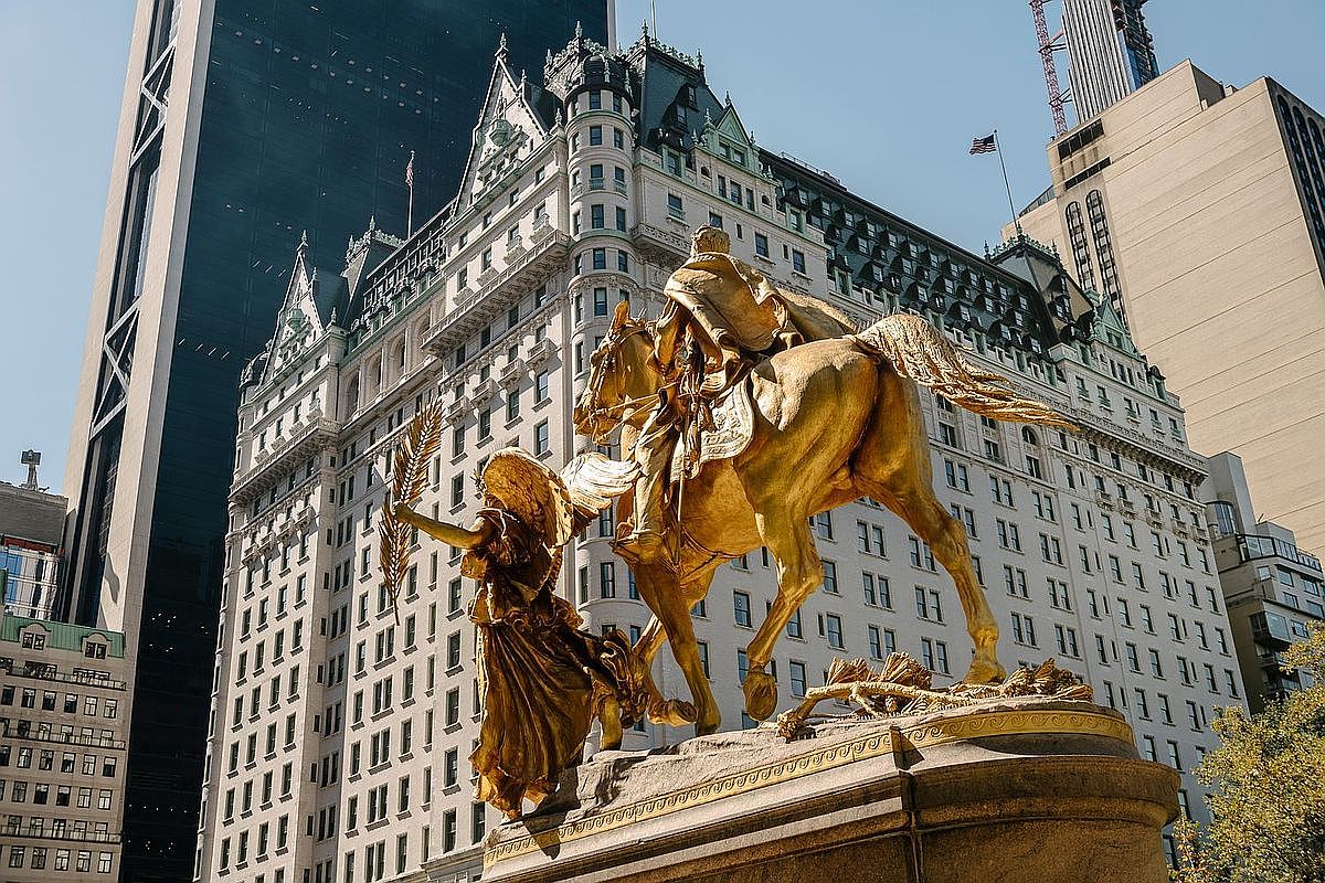 This image captures the iconic General William Tecumseh Sherman Monument, a prominent gilded bronze statue situated in Grand Army Plaza in New York City. The statue features a rider on horseback accompanied by a winged figure, set against the backdrop of the historic Plaza Hotel and surrounding high-rise architecture. The perspective is a low-angle shot that emphasizes the grandeur of the sculpture and the classic Beaux-Arts style of the adjacent building.