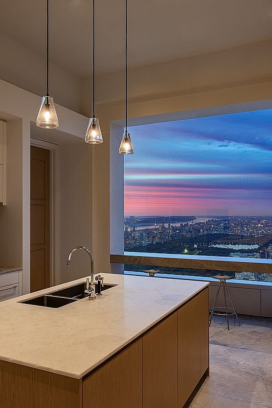 This is a modern kitchen featuring a large island with a white countertop and wooden cabinetry. Three pendant lights hang above the island, and a large window offers a stunning view of a city skyline at dusk. The kitchen is sleek and contemporary, emphasizing the view and natural light.