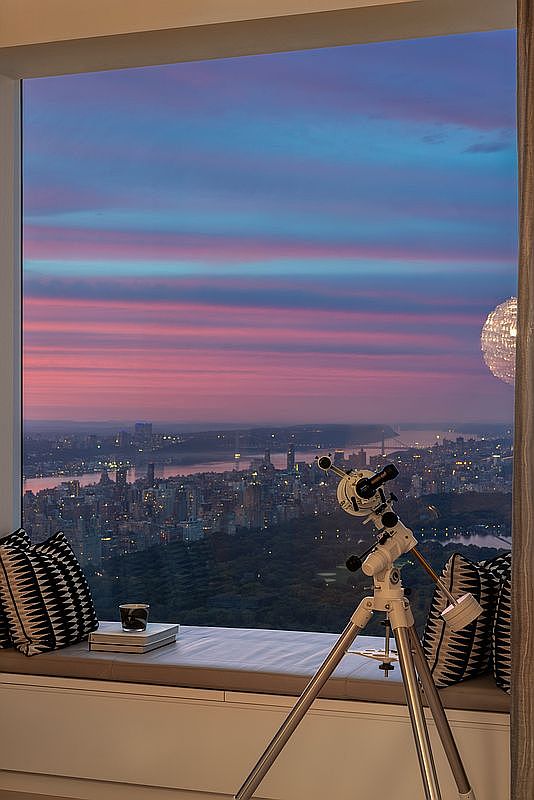 This image showcases a luxurious living room with a breathtaking city view at dusk, framed by a large window. A telescope is positioned near the window, suggesting a focus on the cityscape. The room features comfortable seating with decorative pillows, creating an inviting and sophisticated atmosphere.