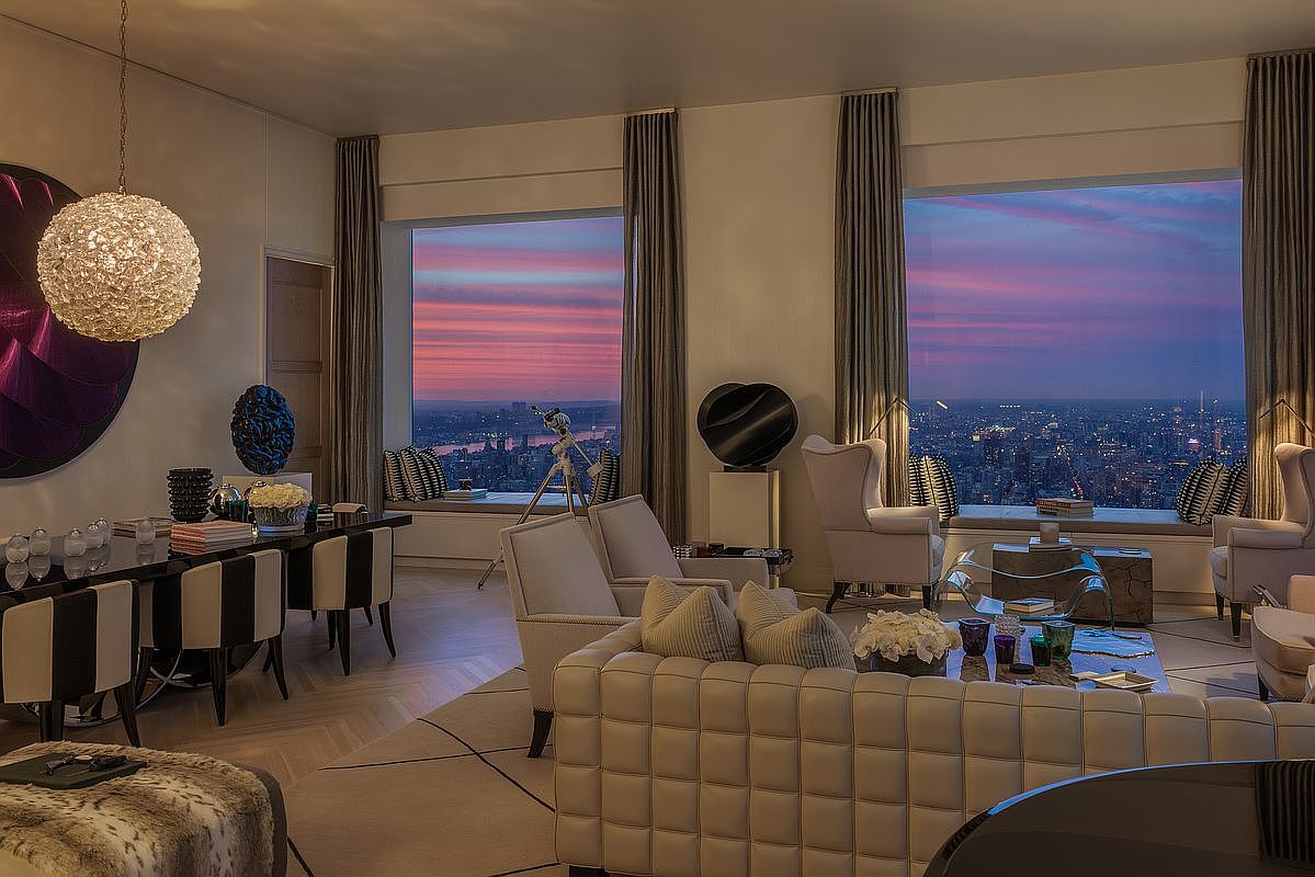 This is a luxurious living room featuring floor-to-ceiling windows with a stunning city view at sunset. The room is furnished with a plush, tufted sofa, elegant armchairs, and a modern dining table with stylish chairs. A large, decorative chandelier hangs above the dining area, adding to the room's sophisticated ambiance.
