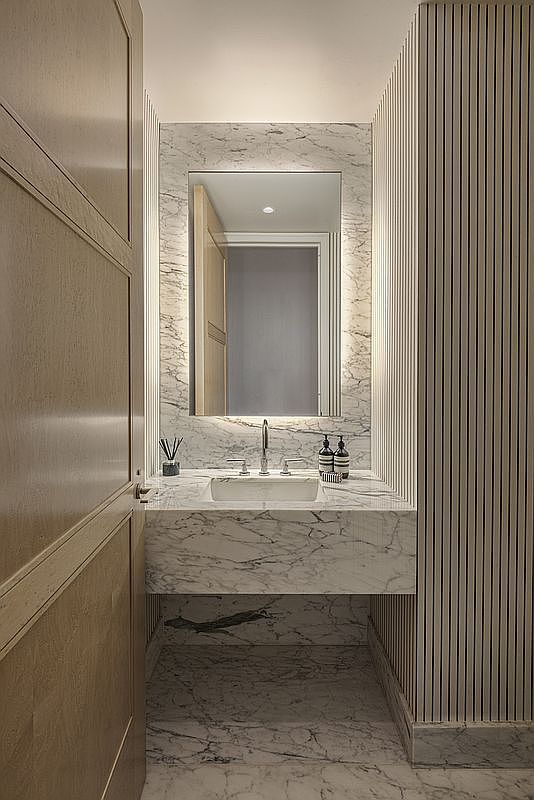 This is a modern guest bathroom featuring a marble countertop and backsplash with a rectangular sink and sleek faucet. A mirror with integrated lighting hangs above the sink, and the walls are adorned with vertical wood paneling. The overall design is clean, elegant, and space-efficient, creating a luxurious feel.