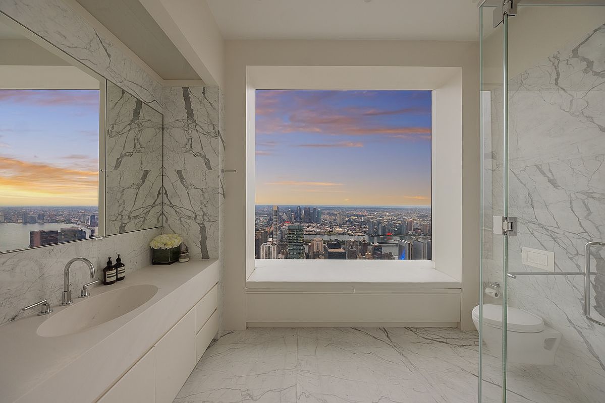 This is a luxurious primary bathroom featuring marble walls and flooring, a modern vanity with an integrated sink, and a large window offering panoramic city views. A glass-enclosed shower and a wall-mounted toilet add to the sleek, contemporary design. The overall impression is one of elegance and sophistication.