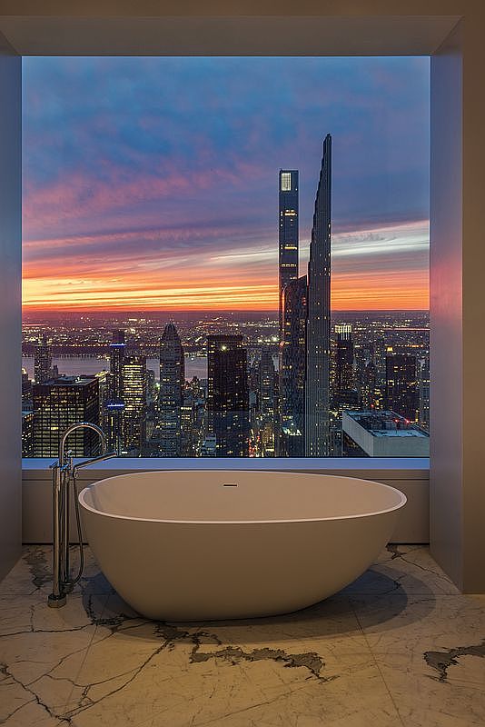 This image showcases a luxurious primary bathroom with a freestanding oval bathtub positioned in front of a large window offering a breathtaking cityscape view at sunset. The marble flooring adds an elegant touch, while the modern faucet complements the sleek design of the tub. The overall impression is one of opulence and relaxation.