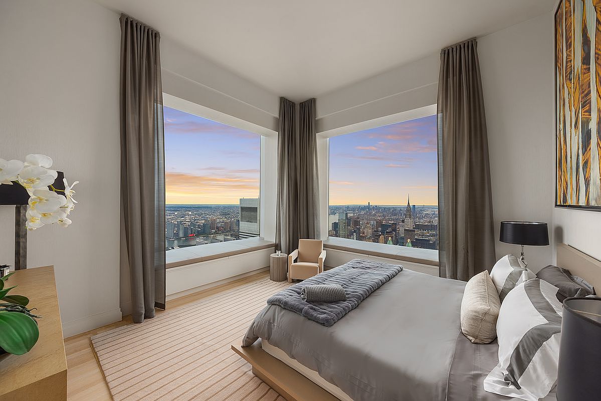 This is a luxurious primary bedroom featuring two large windows with stunning city views at sunset. The room is decorated in neutral tones with a large bed, stylish pillows, and elegant curtains. The overall impression is one of sophistication and comfort, perfect for a high-end real estate listing.