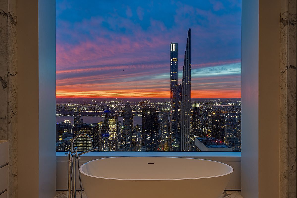 This image showcases a luxurious primary bathroom with a freestanding soaking tub positioned in front of a large window offering a breathtaking cityscape view at sunset. The bathroom features marble accents and modern fixtures, creating a serene and opulent atmosphere. The high-rise buildings and vibrant sky add a dramatic backdrop to the elegant interior.