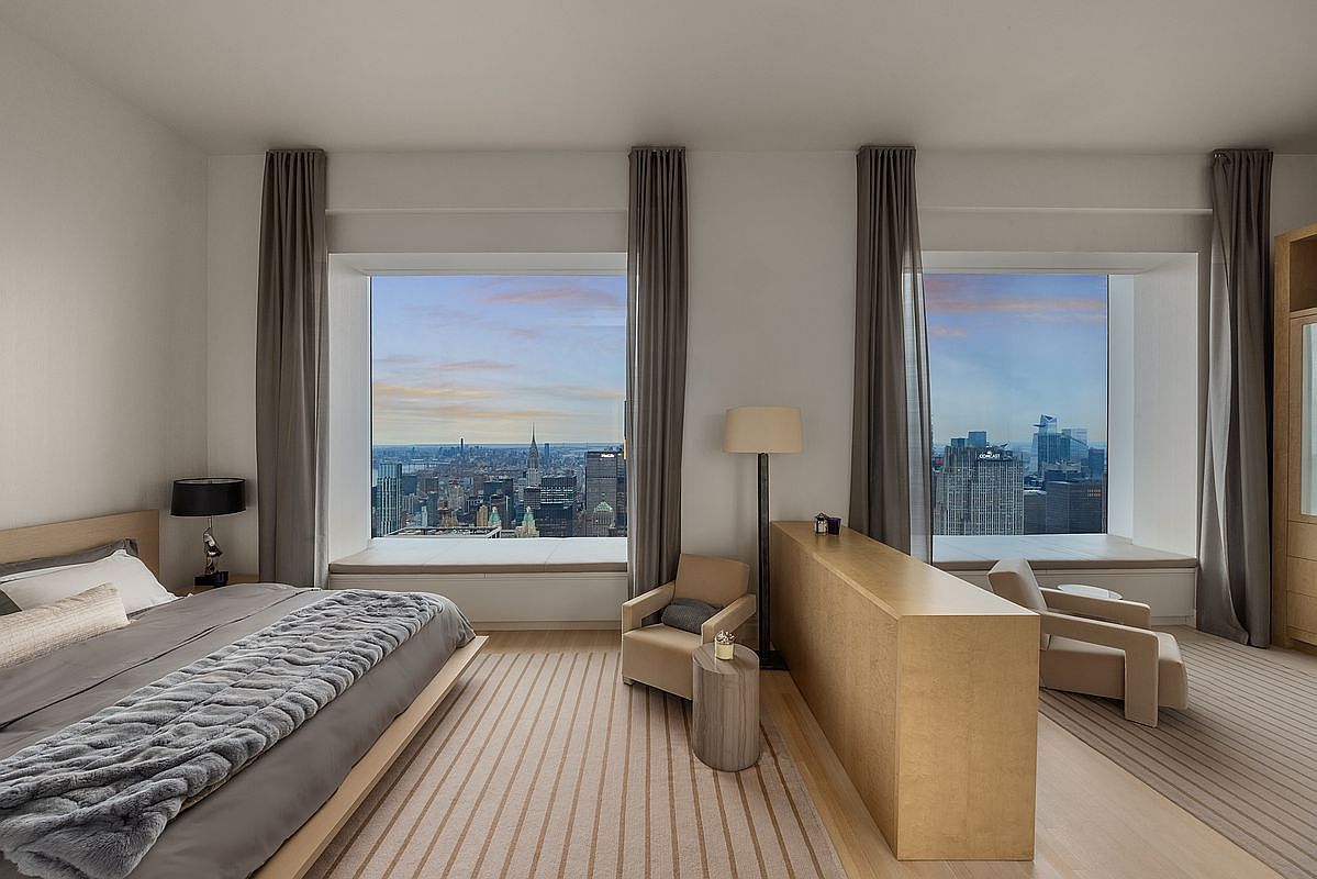 This is a luxurious primary bedroom featuring a large bed with a gray throw, floor-to-ceiling windows offering a stunning city view, and neutral-toned furnishings. The room is bathed in natural light, creating a serene and upscale atmosphere. The design incorporates clean lines and a minimalist aesthetic, emphasizing the breathtaking cityscape.