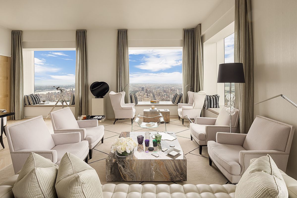 This is a luxurious living room featuring several light-colored armchairs arranged around a marble coffee table. Large windows offer expansive city views, complemented by elegant drapery. The room is decorated in a neutral palette, creating a sophisticated and inviting atmosphere.