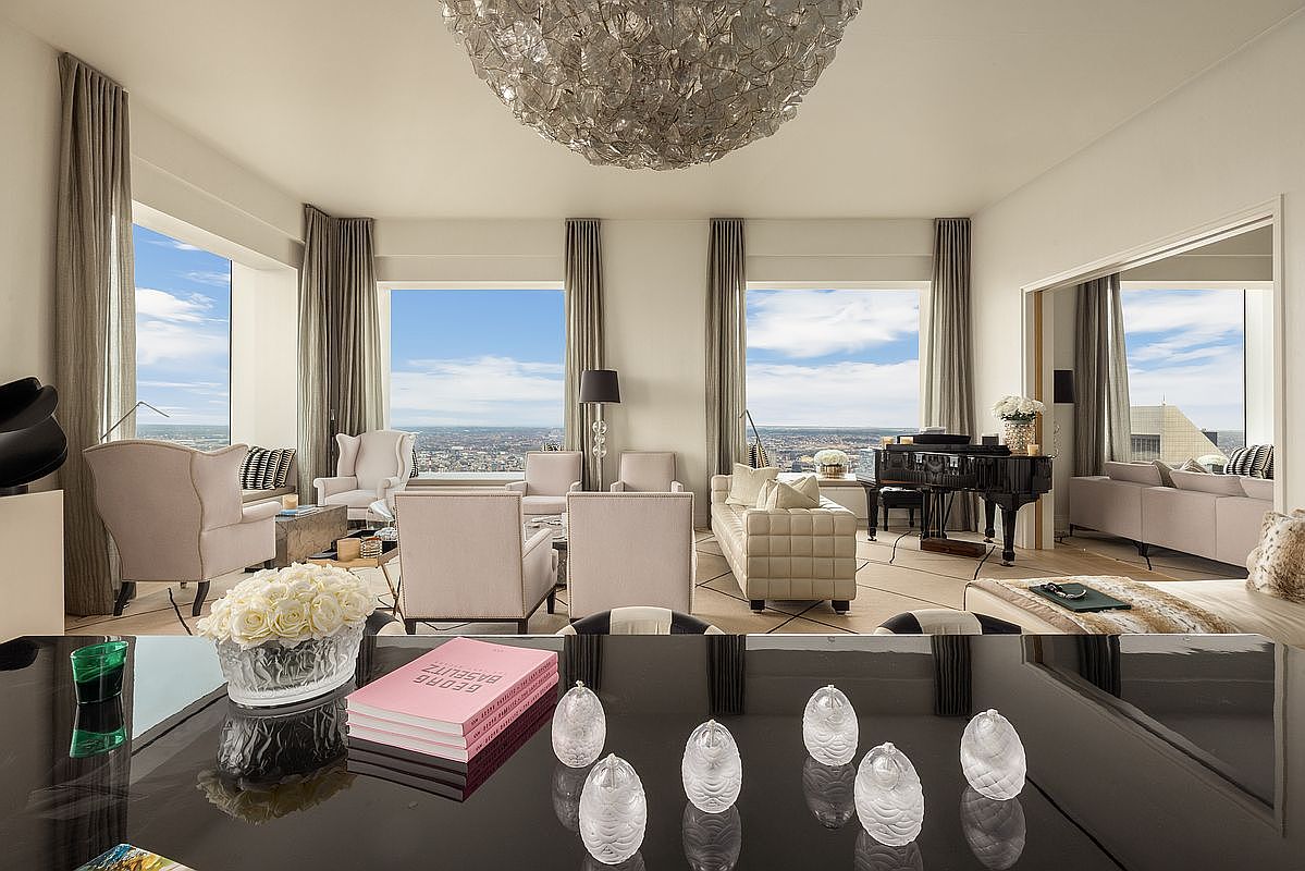 This is a luxurious living room featuring high ceilings, large windows with city views, and elegant furnishings. The room includes comfortable seating arrangements, a grand piano, and a striking chandelier, creating a sophisticated and inviting atmosphere. The perspective is from a low angle, emphasizing the room's grandeur and depth.