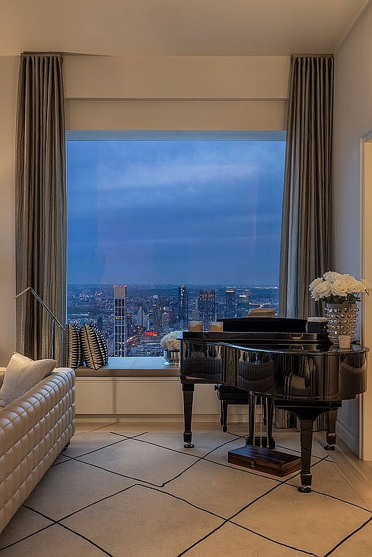 This interior shot showcases a luxurious living room with a grand piano positioned in front of a large window offering a stunning city view at dusk. The room features elegant curtains, a stylish sofa, and a patterned rug, creating a sophisticated and inviting atmosphere. The perspective is from a medium distance, capturing the room's depth and the impressive cityscape beyond.