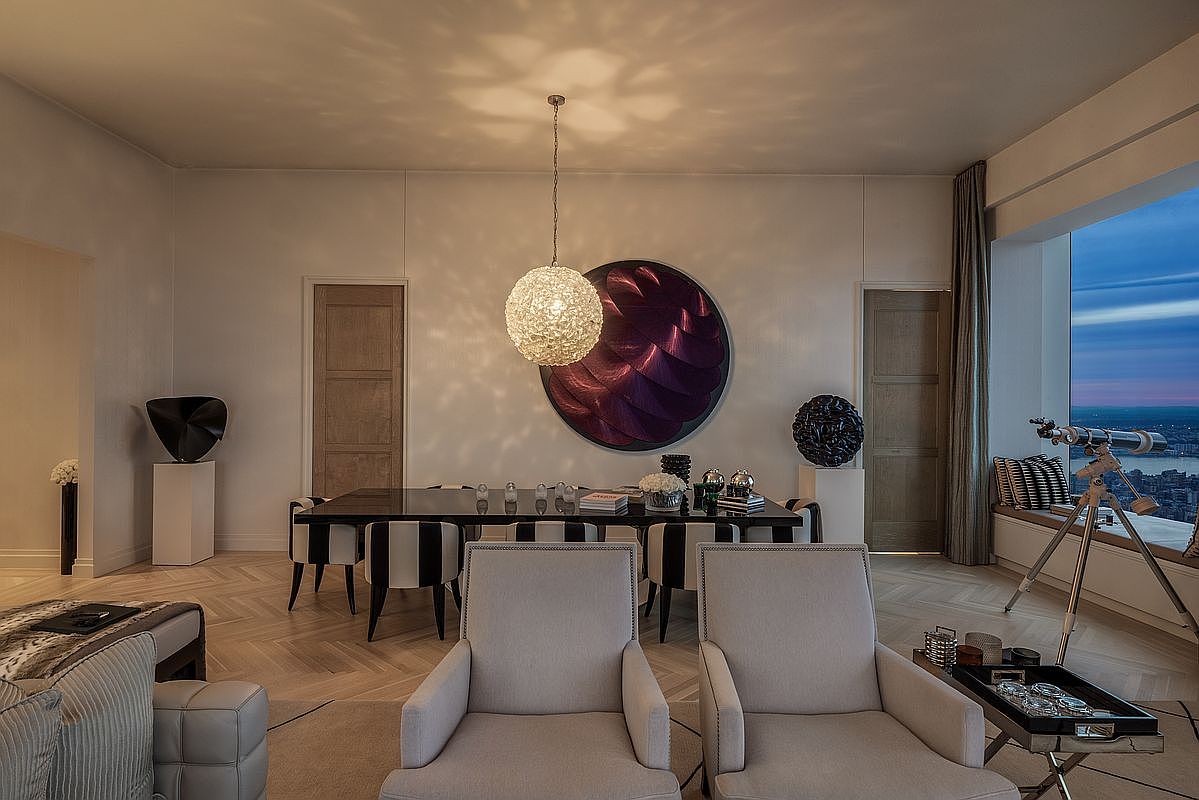 This is an elegant dining room featuring a long, dark wood table with striped chairs, illuminated by a modern, spherical pendant light. A large, circular artwork adorns the wall, adding a touch of sophistication. The room also includes two armchairs in the foreground and a telescope by the window, suggesting a luxurious and refined living space.