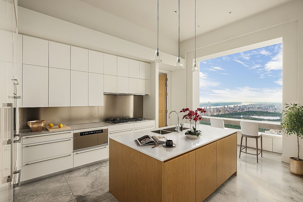 This is a bright, modern kitchen featuring sleek white cabinetry and a large island with a light wood finish and white countertop. Stainless steel appliances and pendant lighting add to the contemporary feel. A large window offers a stunning city view, enhancing the luxurious ambiance of the space.