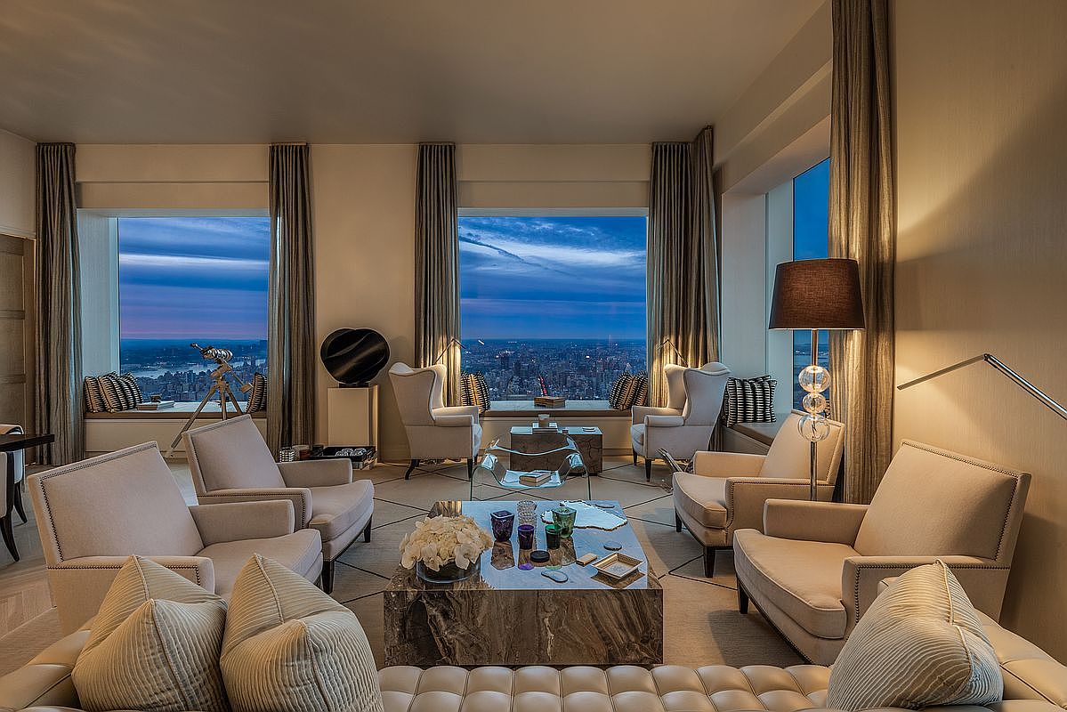 This is a luxurious living room featuring floor-to-ceiling windows with a stunning city view at dusk. The room is furnished with plush armchairs, a marble coffee table, and a comfortable sofa, all arranged on a patterned rug. The neutral color palette and elegant lighting create a sophisticated and inviting atmosphere, perfect for relaxation and entertaining.