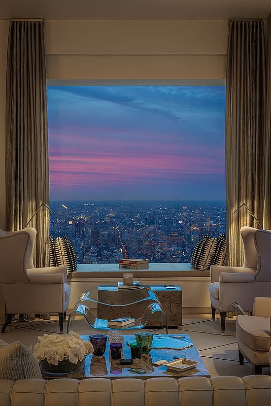 This is a luxurious living room featuring a breathtaking cityscape view through a large window framed by elegant curtains. The room is furnished with plush armchairs, a glass coffee table, and a comfortable sofa, creating a sophisticated and inviting atmosphere. The overall impression is one of high-end urban living with a focus on comfort and style.