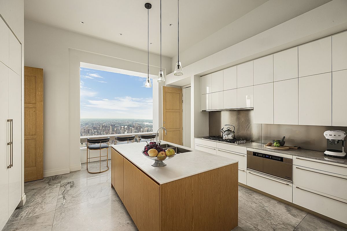 This is a bright, modern kitchen featuring sleek white cabinetry and a contrasting wood-paneled island with a white countertop. Stainless steel appliances and pendant lighting add to the contemporary aesthetic. A large window offers a stunning city view, creating a luxurious and airy atmosphere.