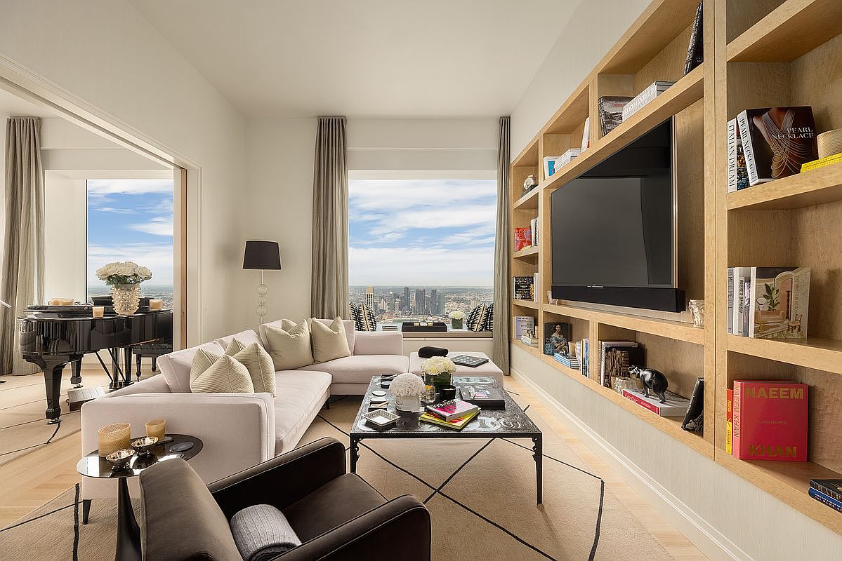 This is a luxurious living room featuring a plush sectional sofa, a dark wood coffee table, and a built-in bookshelf with a large television. A grand piano sits near a large window offering a stunning city view. The room is decorated in neutral tones with pops of color from books and accessories, creating an elegant and inviting atmosphere.