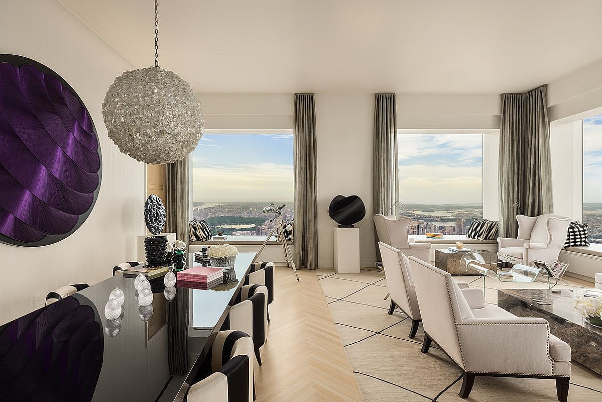 This is an interior shot showcasing a luxurious dining and living area with stunning city views. The dining area features a sleek, black dining table surrounded by stylish chairs, complemented by a modern chandelier and a large, abstract purple artwork. The living area includes comfortable armchairs and a unique coffee table, all set against a backdrop of expansive windows offering panoramic vistas.