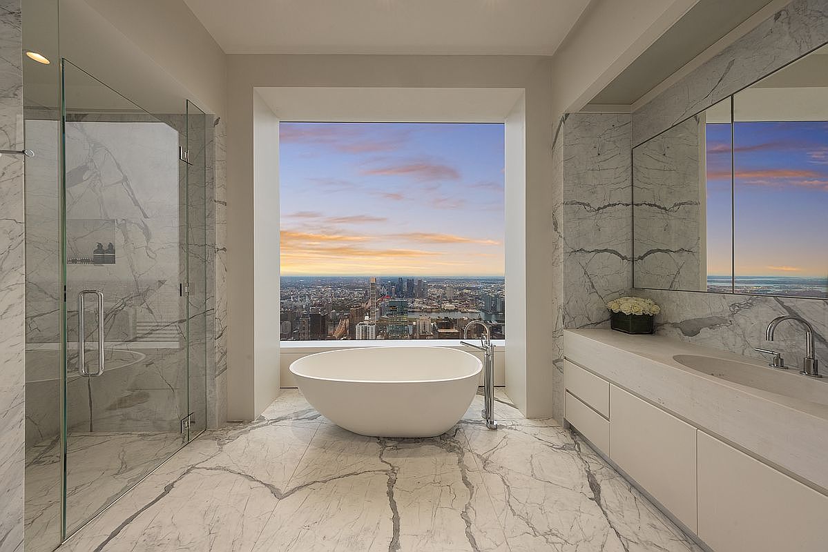 This is a luxurious primary bathroom featuring marble walls and flooring, a freestanding soaking tub positioned in front of a large window with a city view, and a glass-enclosed shower. A modern vanity with integrated sink and storage completes the space. The overall impression is one of opulence and relaxation.