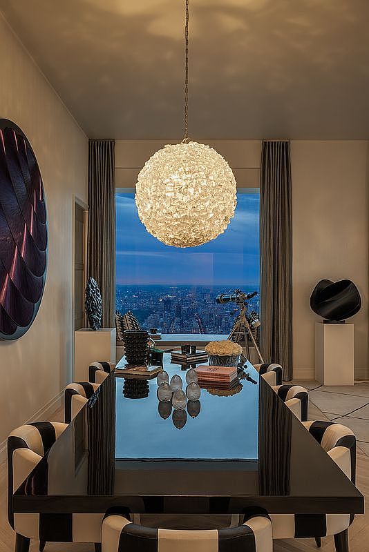 This is an interior shot of a luxurious dining room featuring a glossy black dining table surrounded by black and white striped chairs. A large, spherical chandelier hangs above the table, and floor-to-ceiling windows offer a stunning city view at dusk. The room is decorated with modern art pieces and sculptures, creating a sophisticated and elegant atmosphere.
