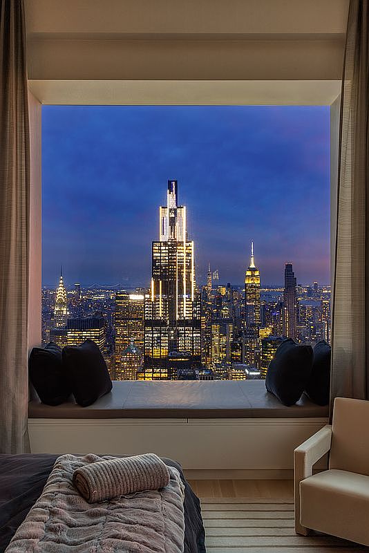 This is a luxurious primary bedroom featuring a stunning cityscape view through a large window. The room is elegantly furnished with a comfortable bed, a cozy window seat with pillows, and a stylish armchair. The overall impression is one of sophistication and urban tranquility, highlighting the premium location and upscale living.