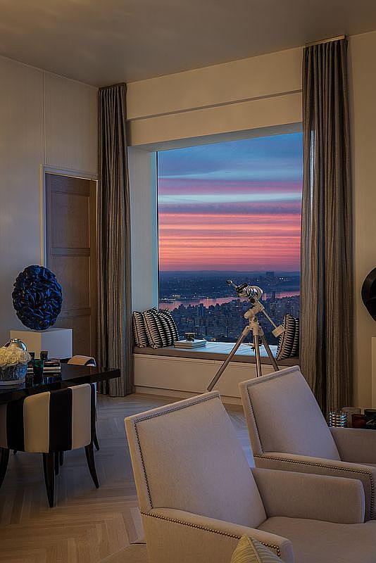 This is an interior shot of a luxurious living room featuring a stunning city view through a large window at sunset. The room is elegantly furnished with comfortable armchairs, a dining table with stylish chairs, and a telescope positioned to capture the cityscape. The overall impression is one of sophistication and urban living.