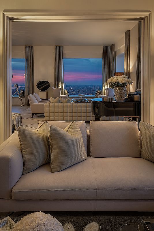 This image showcases a luxurious living room with a sophisticated and elegant design. The room features plush sofas with decorative pillows, a dark piano, and large windows offering a stunning city view at dusk. The neutral color palette and high-end furnishings create a serene and upscale atmosphere, perfect for a discerning buyer.