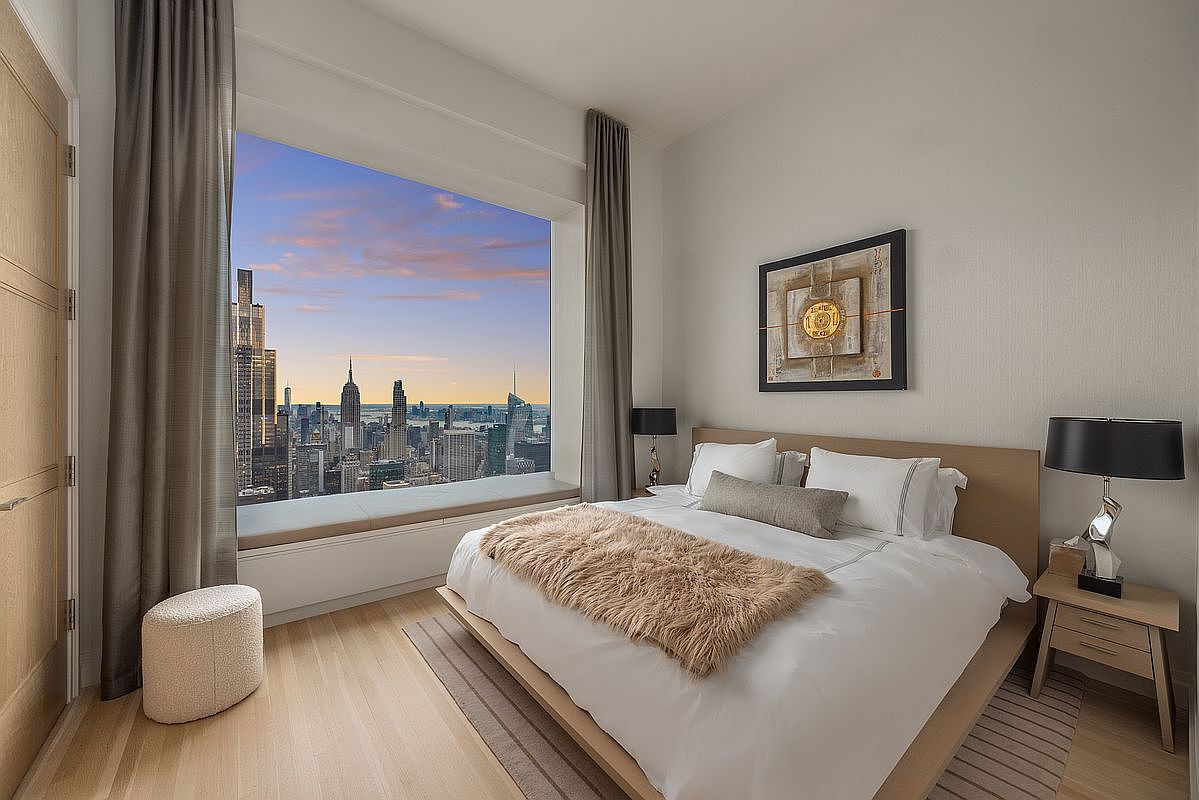 This is a luxurious primary bedroom featuring a king-size bed with white linens and a faux fur throw. A large window offers a stunning city view, complemented by neutral-toned curtains. The room is decorated with modern art and minimalist furniture, creating a serene and upscale atmosphere.
