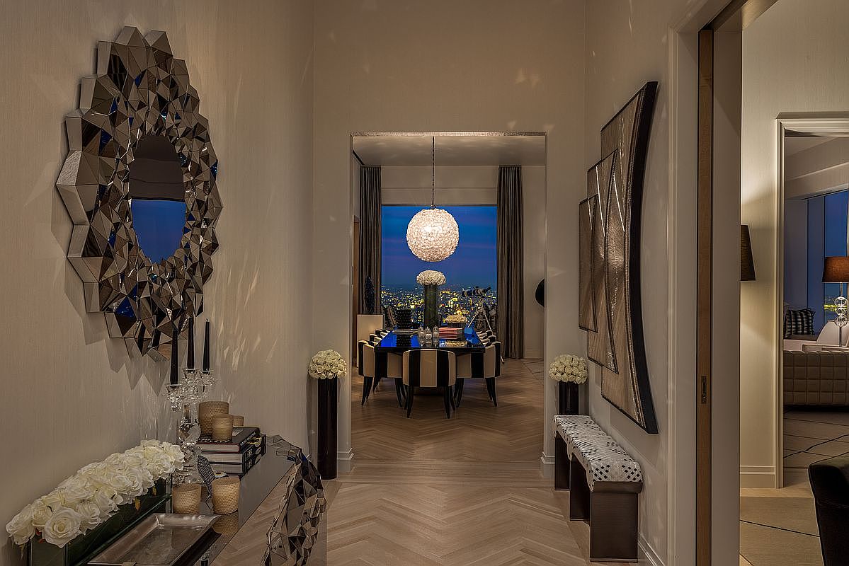This interior shot showcases a luxurious hallway leading to a dining area with a stunning city view. The hallway features a large, geometric mirror, a mirrored console table adorned with flowers, and an elegant piece of artwork complemented by a stylish bench. The herringbone wood flooring adds warmth and sophistication to the space, creating an inviting and upscale ambiance.