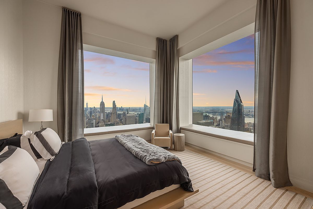 This primary bedroom boasts stunning city views through expansive windows, complemented by elegant gray curtains. The room features a large bed with dark bedding and striped pillows, a neutral-toned chair, and a light-colored striped rug, creating a luxurious and serene atmosphere. The natural light and sophisticated design make this a desirable space.