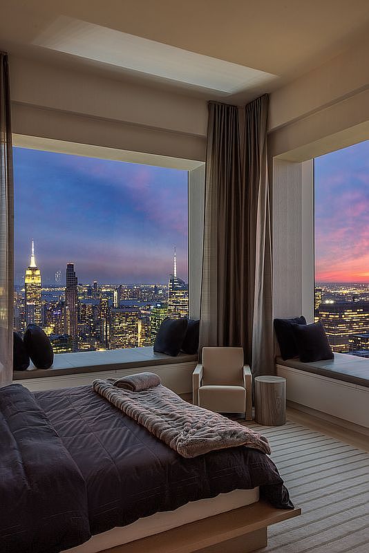 This is a luxurious primary bedroom featuring a large bed with dark bedding and a textured throw. Floor-to-ceiling windows offer stunning city views at dusk, with the Empire State Building visible. The room includes a seating area with a chair and a small side table, and the overall design is modern and elegant.