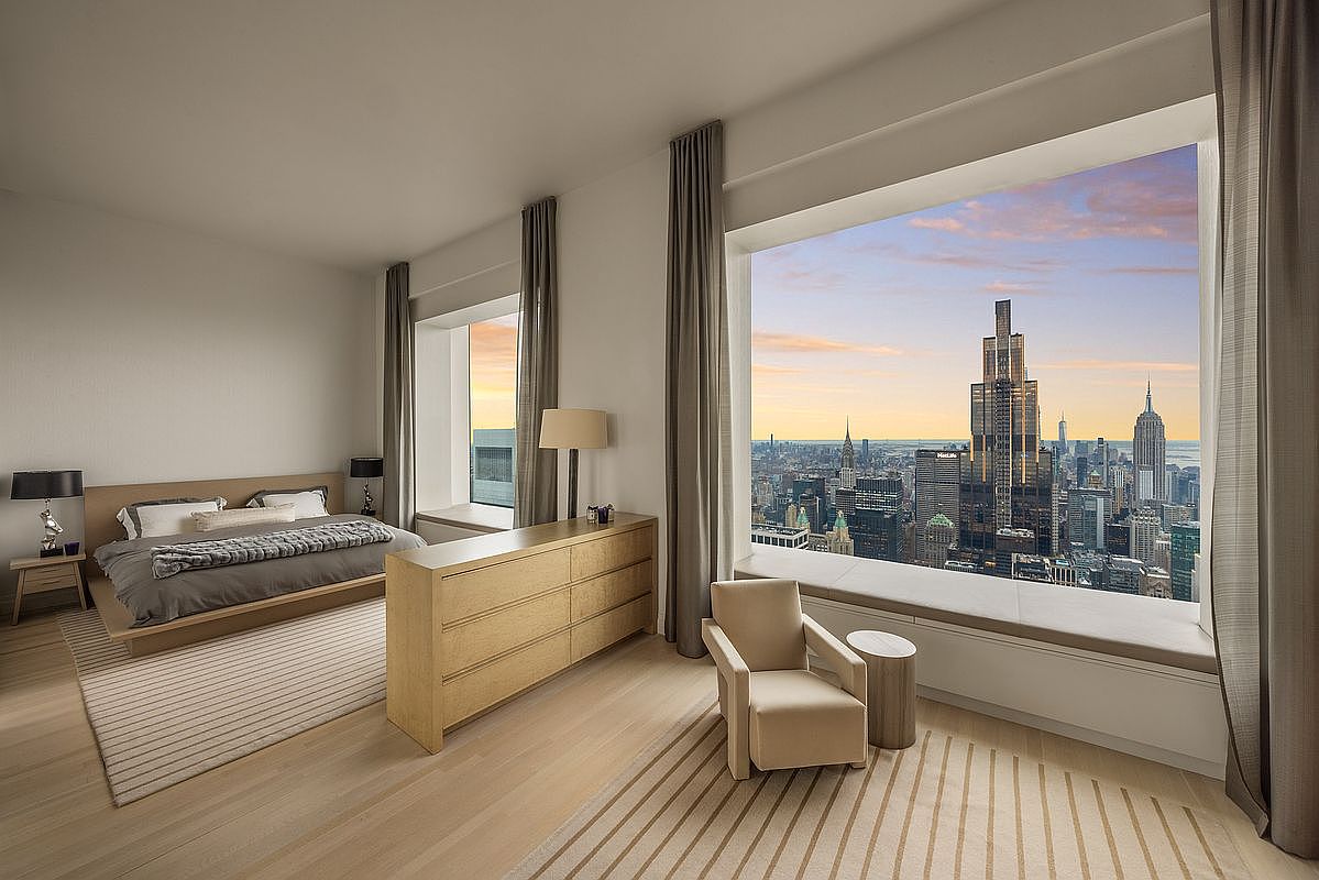 This is a luxurious primary bedroom featuring a large window with a stunning city view. The room is decorated in neutral tones with a modern bed, a wooden dresser, and a comfortable armchair. The overall impression is one of elegance and tranquility, enhanced by the natural light and expansive cityscape.