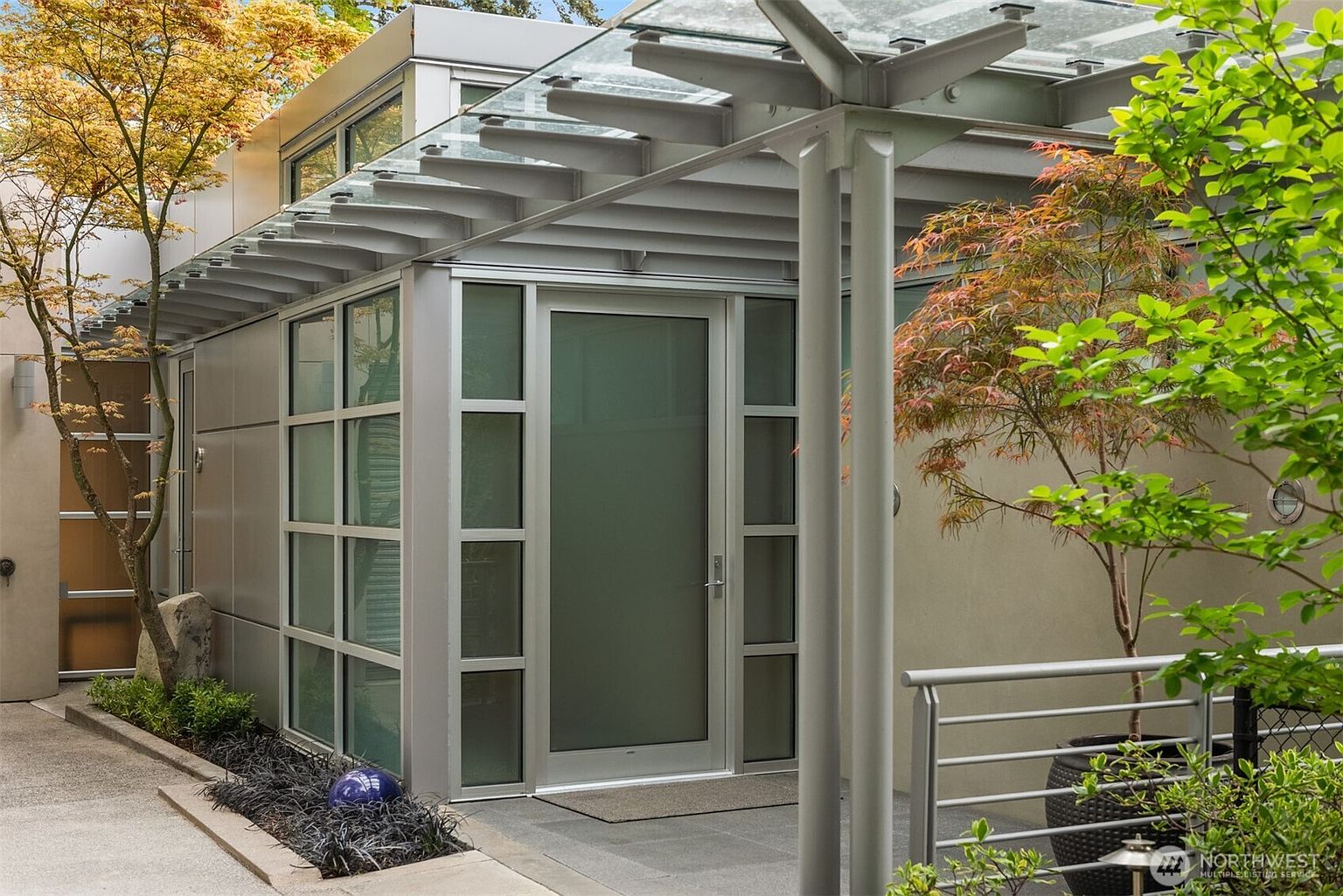 This modern entryway features a sleek glass-paneled door framed by frosted glass sidelights and a contemporary metal structure. A glass canopy extends over the entrance, supported by metallic columns, creating a sophisticated and architectural aesthetic. The surrounding area is landscaped with a small tree and dark ground cover, enhancing the clean, minimalist design of the home's exterior.
