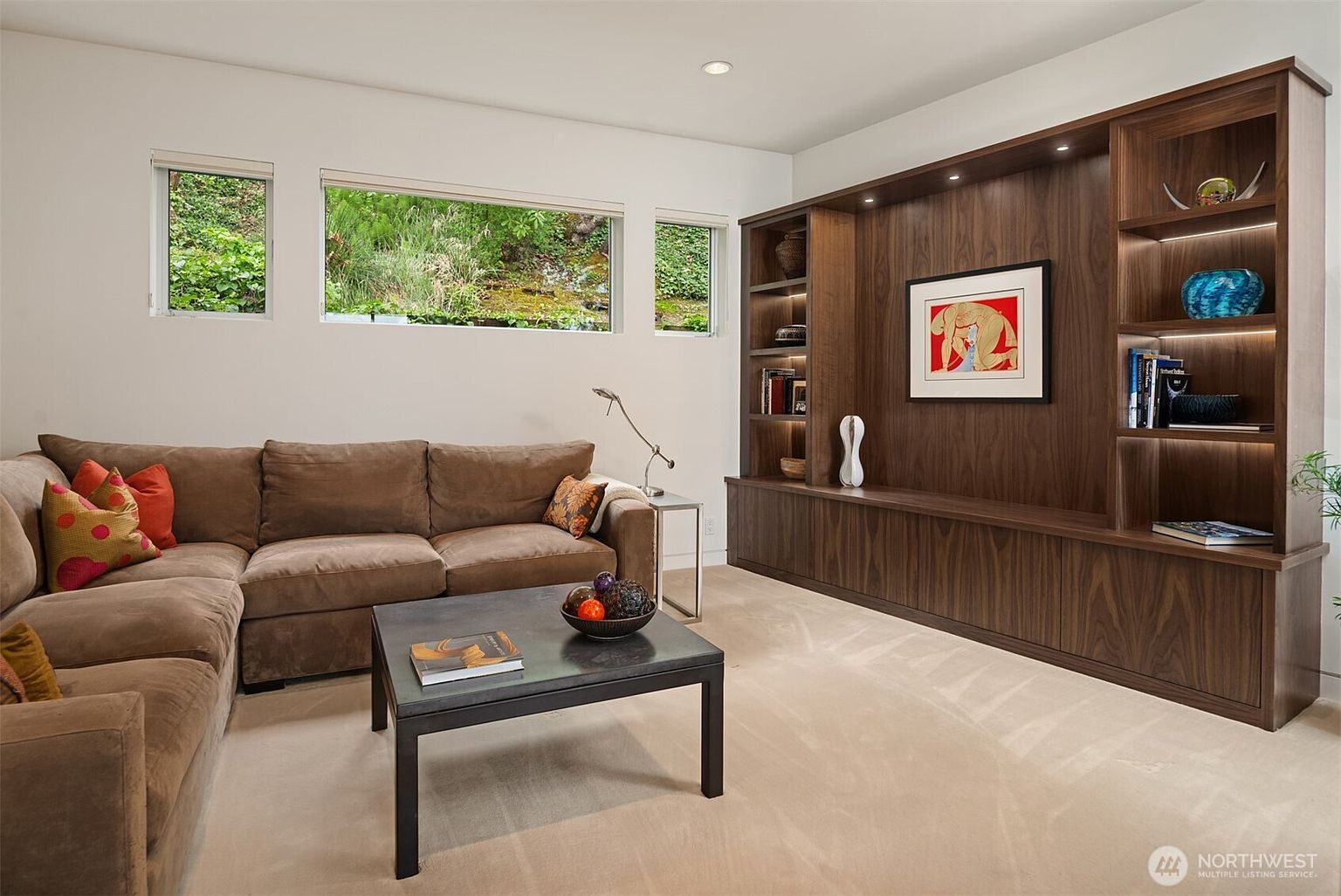 This contemporary living room features a large, plush brown sectional sofa arranged around a dark metal-framed coffee table. A prominent, custom-built dark wood media unit with integrated lighting serves as the focal point, displaying decorative items and artwork. Three horizontal windows along the wall provide natural light and a view of the lush greenery outside, creating a serene and inviting atmosphere.