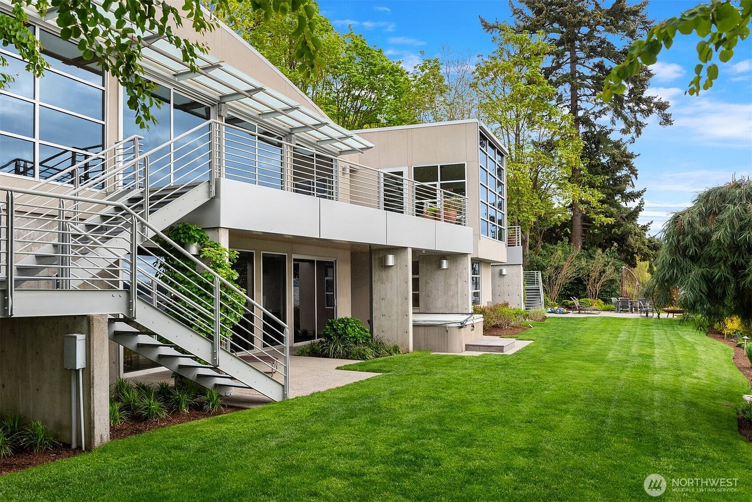 This modern architectural home features a striking multi-level design with concrete structural elements, metal railings, and expansive glass walls. A prominent exterior staircase connects the upper deck to the manicured lawn, which leads toward a lush, tree-lined backyard. The scene captures a serene outdoor living space, complete with a built-in spa on the lower patio, emphasizing a seamless blend of contemporary design and natural surroundings.