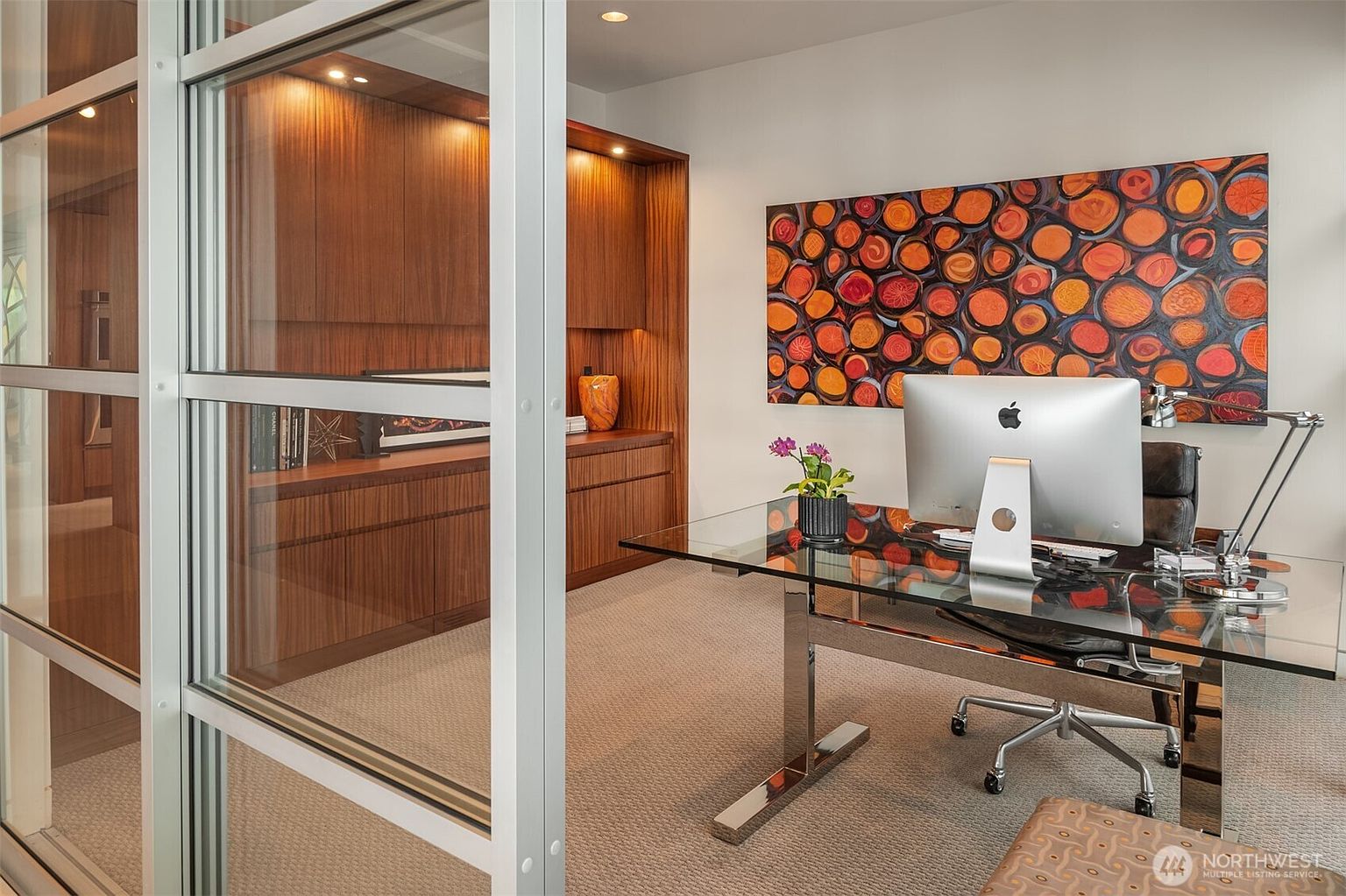 This modern home office features a sleek glass-topped desk and a comfortable leather chair, positioned in front of a vibrant, abstract wall art piece. The room is anchored by extensive custom wood cabinetry that provides ample storage and display space, complemented by warm recessed lighting. A glass-paneled partition wall frames the view, creating a professional and sophisticated workspace.
